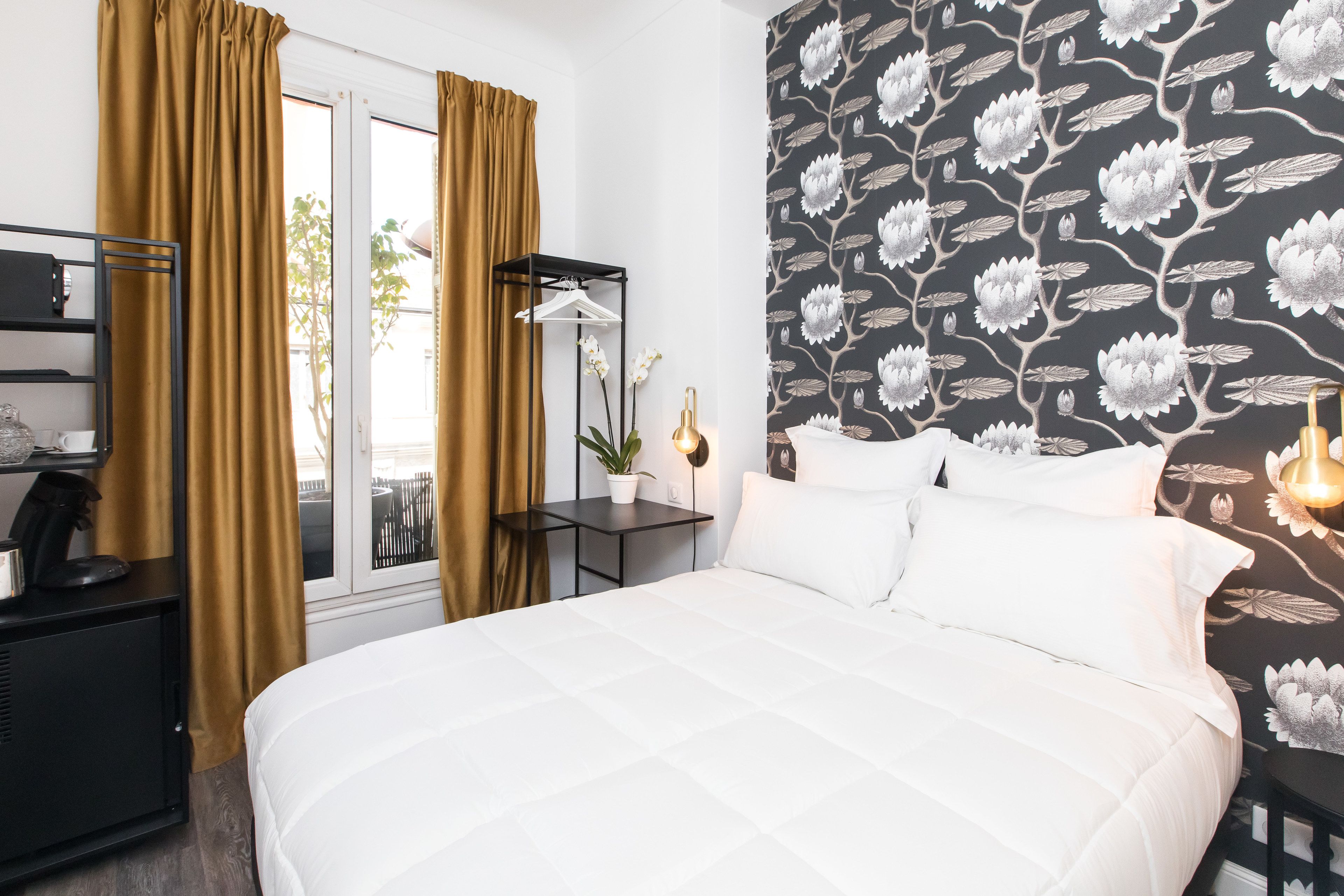 Ein Hotelzimmer mit Bett, Nachttisch, Fenster mit Vorhängen, Wand mit Blumenmuster und einem kleinen Tisch mit Pflanze.