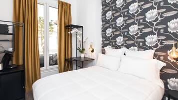 Ein Hotelzimmer mit Bett, Nachttisch, Fenster mit Vorhängen, Wand mit Blumenmuster und einem kleinen Tisch mit Pflanze.
