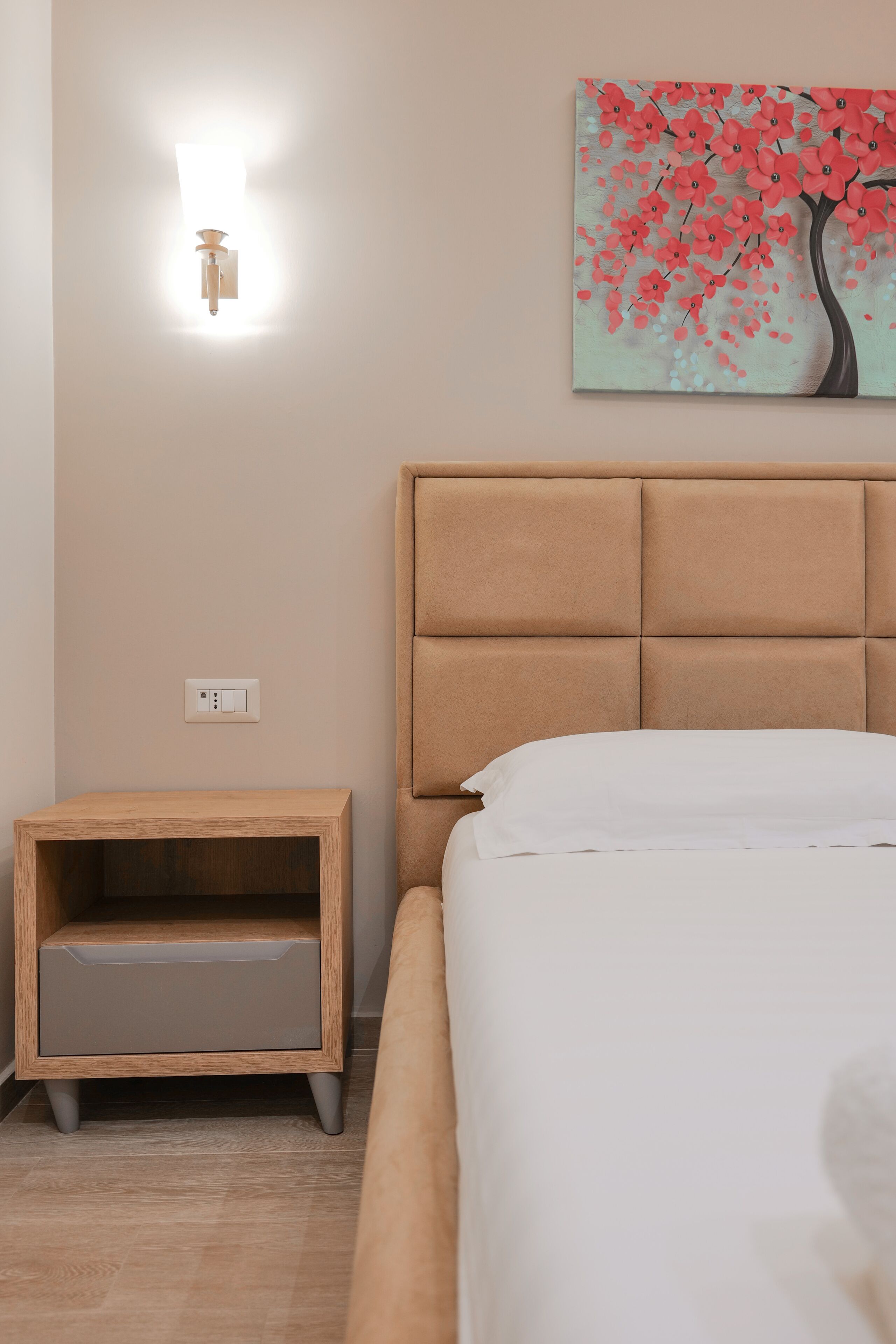 A bedroom with a bed, a nightstand, a wall-mounted light, and a painting of a tree with pink flowers.