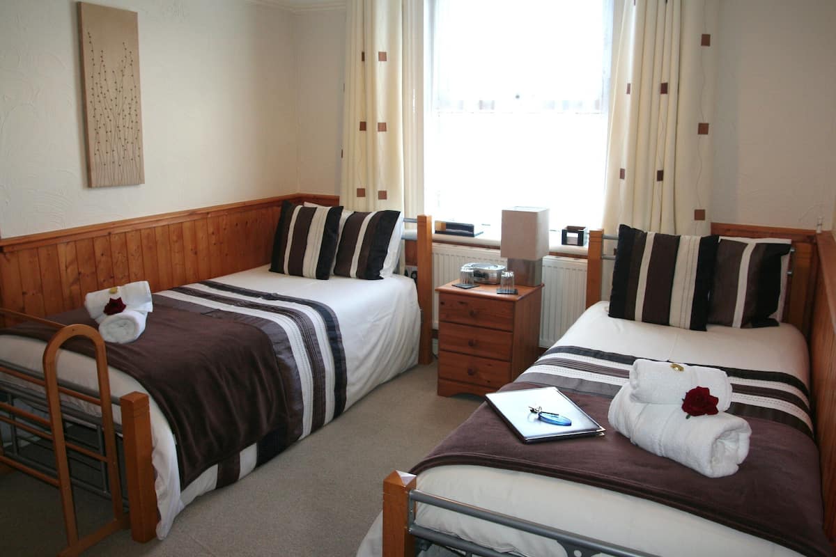 Een hotelkamer met twee eenpersoonsbedden, houten hoofdborden, een klein houten nachtkastje, een radiator en een raam met gordijnen.
