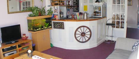 Una sala de estar con televisor, mesa de centro de madera, sofá, acuario, área de cocina con refrigerador y barra.
