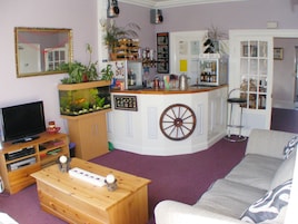 Una sala de estar con televisor, mesa de centro de madera, sofá, acuario, área de cocina con refrigerador y barra.