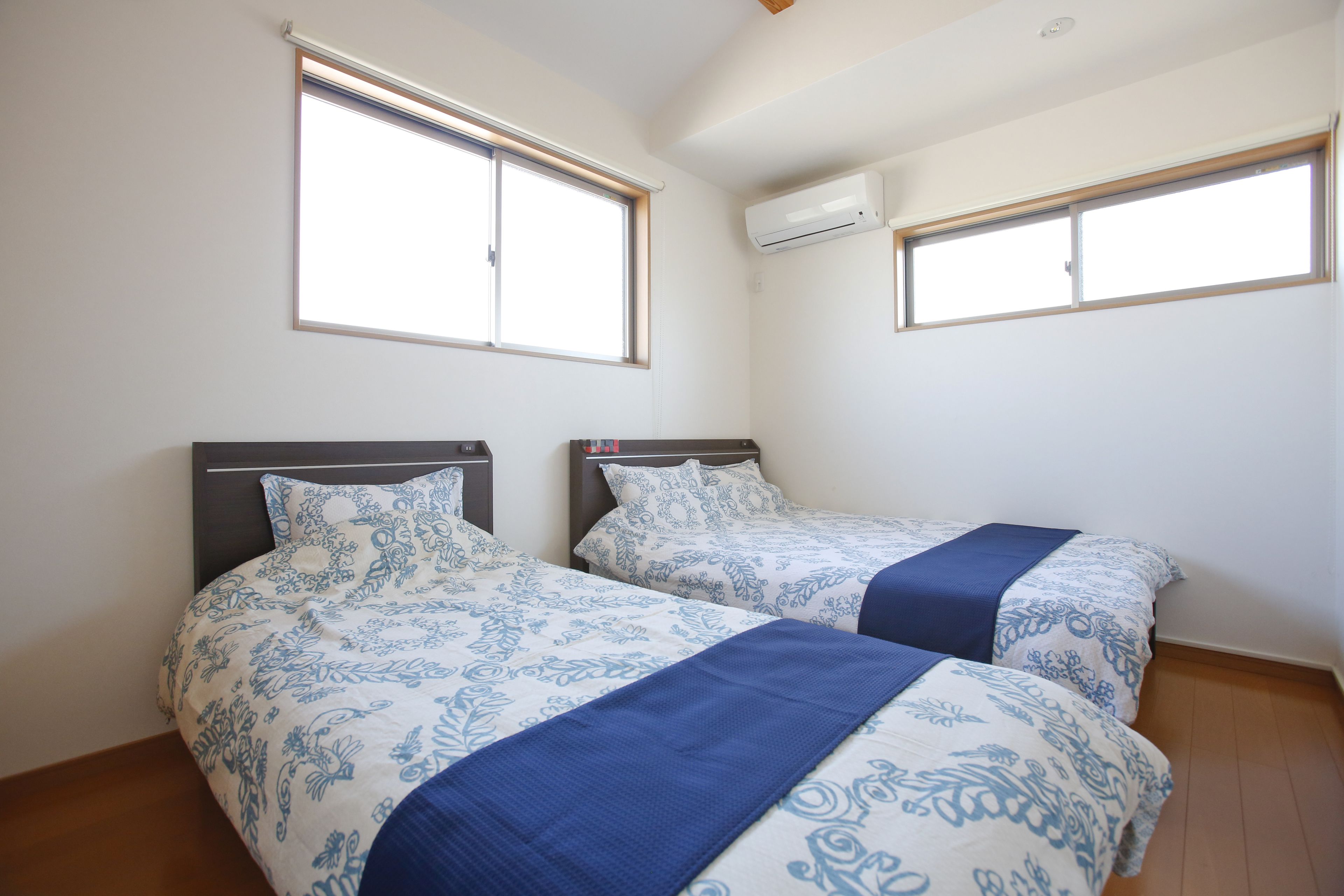 Une chambre avec deux lits, chacun doté d’une tête de lit et d’une fenêtre. Un appareil de climatisation est fixé au mur.