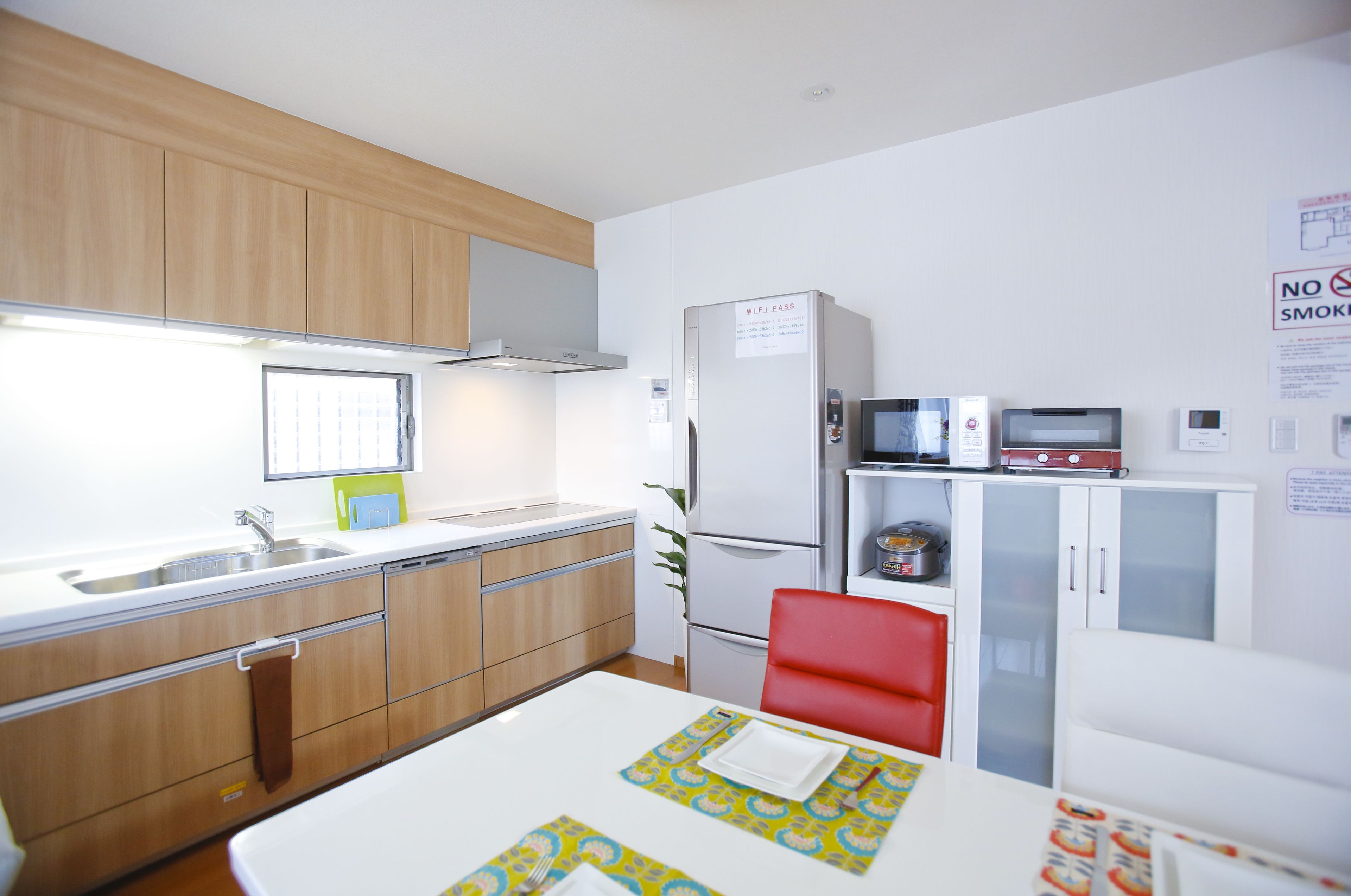 A compact kitchen with wooden cabinets, a white countertop, a microwave, a refrigerator, and a dining area with a red chair and a white table.