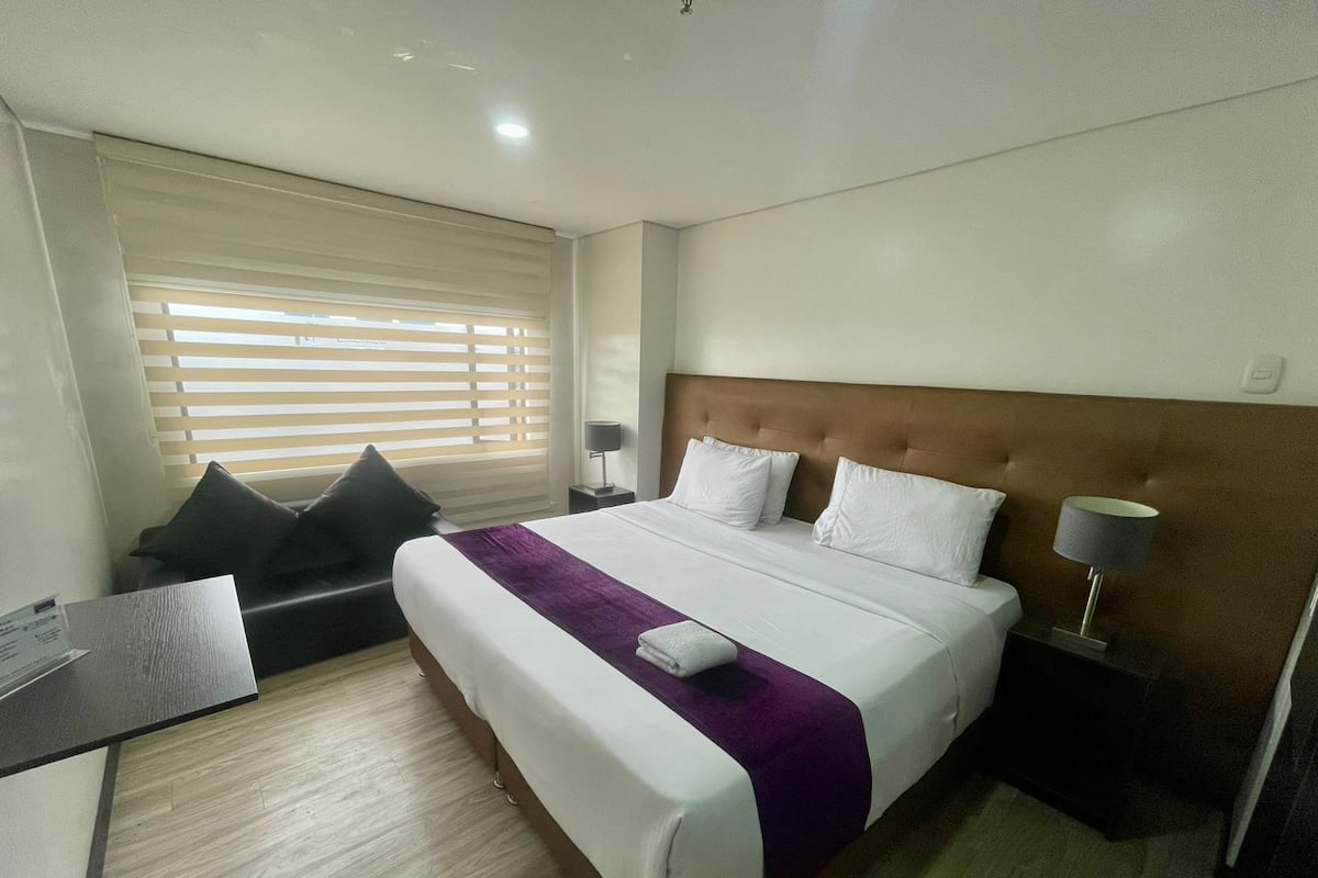 A hotel room with a bed, a desk, a chair, a window with blinds, and a nightstand with a lamp.