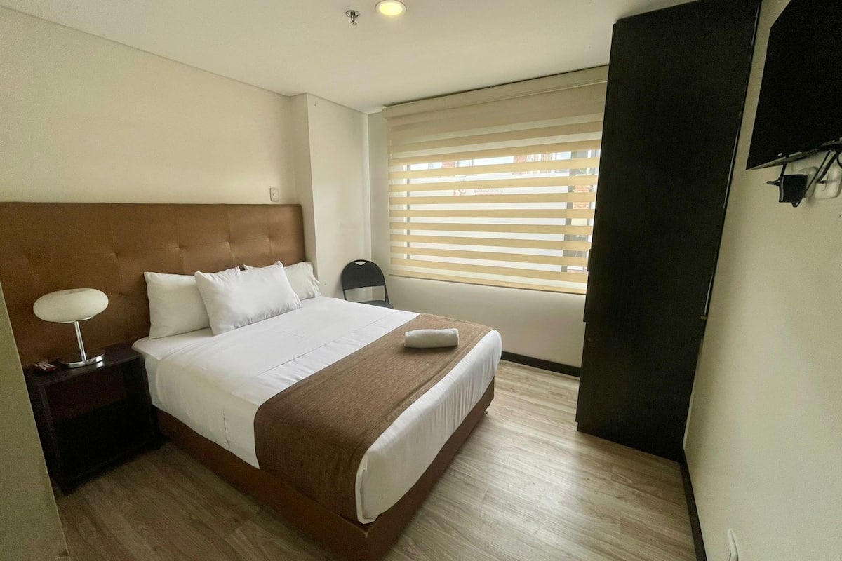 A hotel room with a large bed, a nightstand, a chair, and a window with blinds.