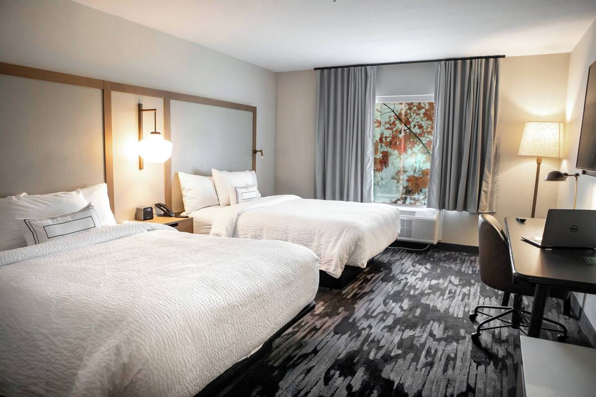 A hotel room with two beds, a desk with a laptop, a desk lamp, a phone, and a window with curtains.