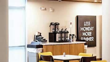 A modern cafe with yellow chairs and tables, a coffee machine, and a motivational poster on the wall.