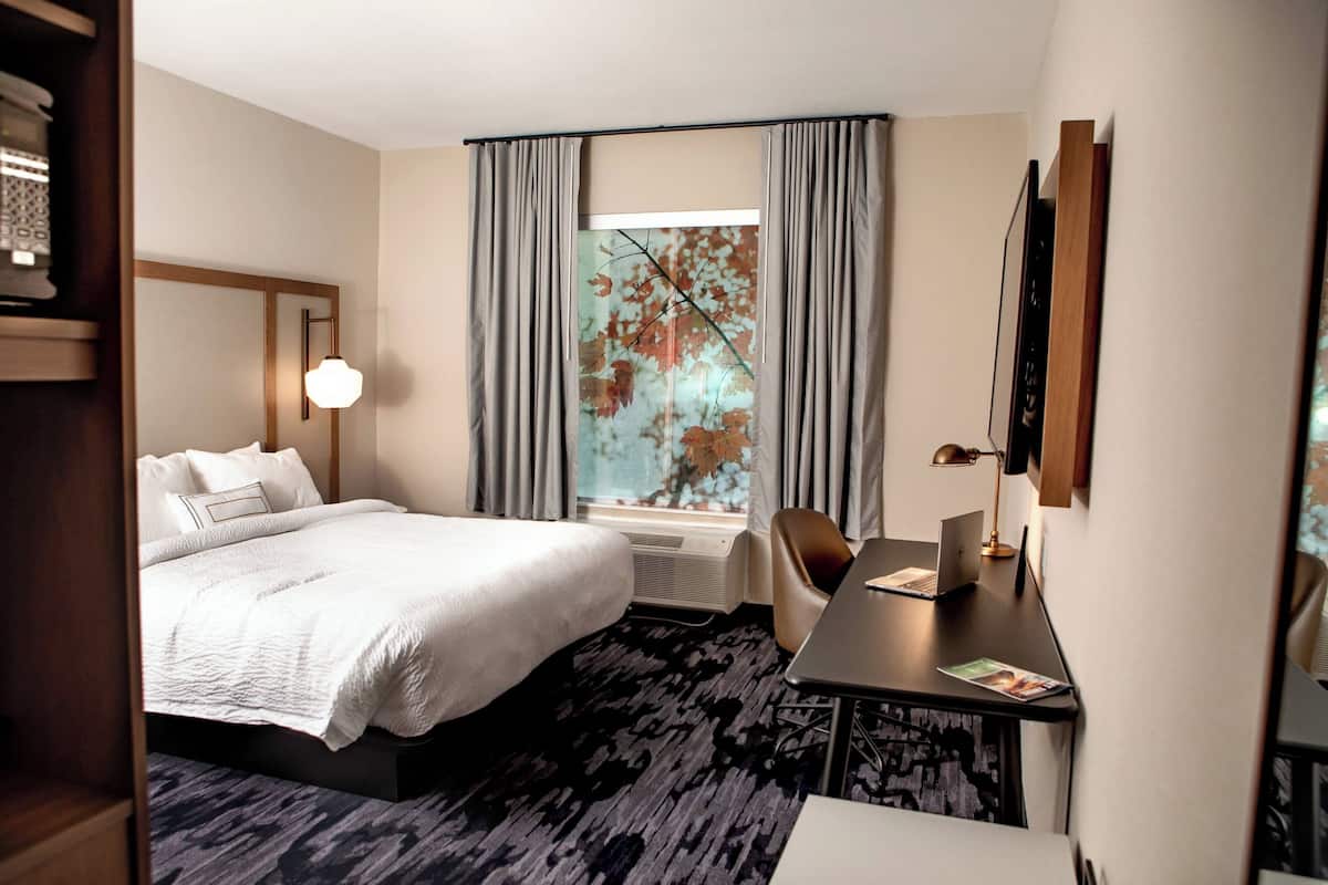 A hotel room with a bed, a desk with a laptop, a chair, a window with curtains, and a wall-mounted TV.