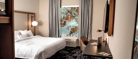 A hotel room with a bed, a desk with a laptop, a chair, a window with curtains, and a wall-mounted TV.