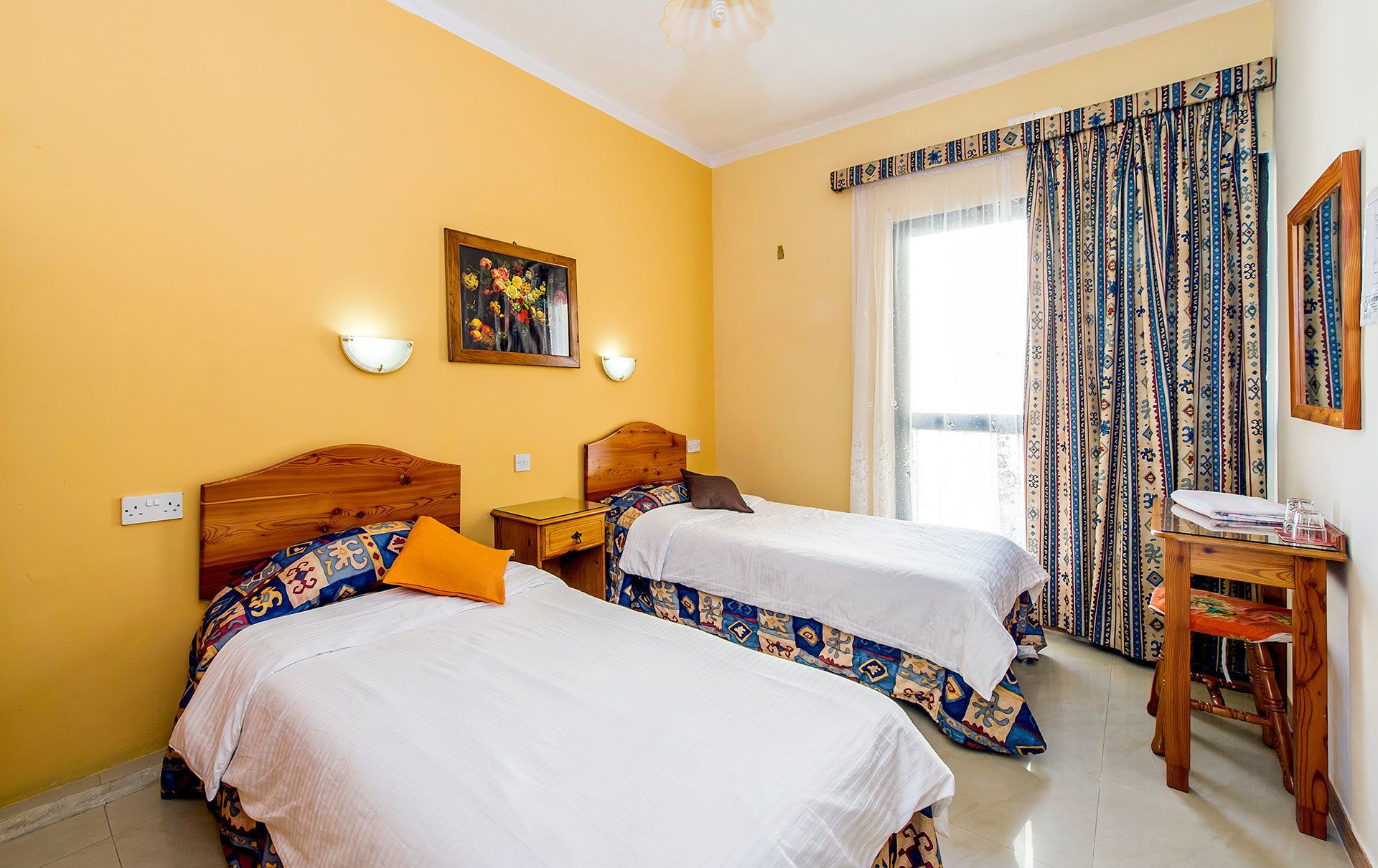 Habitación de hotel con dos camas, una mesita de noche de madera, una pequeña mesa y una ventana con cortinas estampadas.