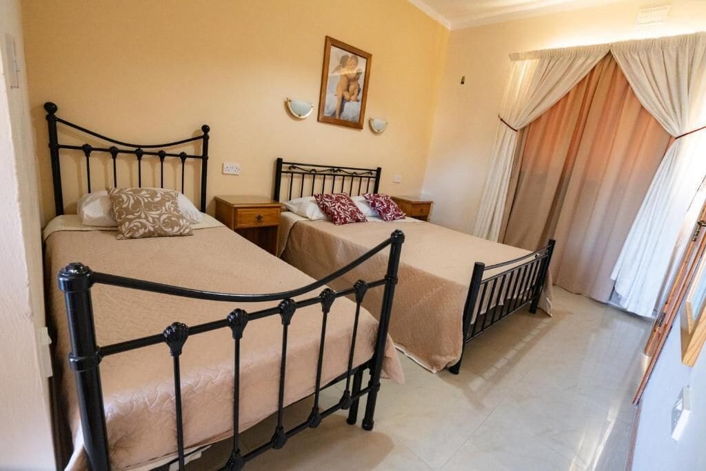 Une chambre avec deux lits, une table de chevet, une lampe fixée au mur et une fenêtre avec des rideaux.