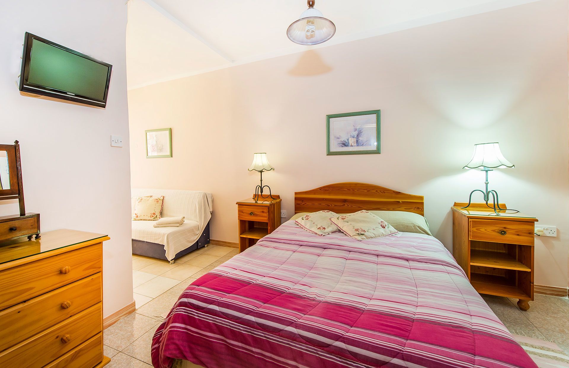 A bedroom with a bed, two bedside tables with lamps, a dresser, a chair, and a television on the wall.