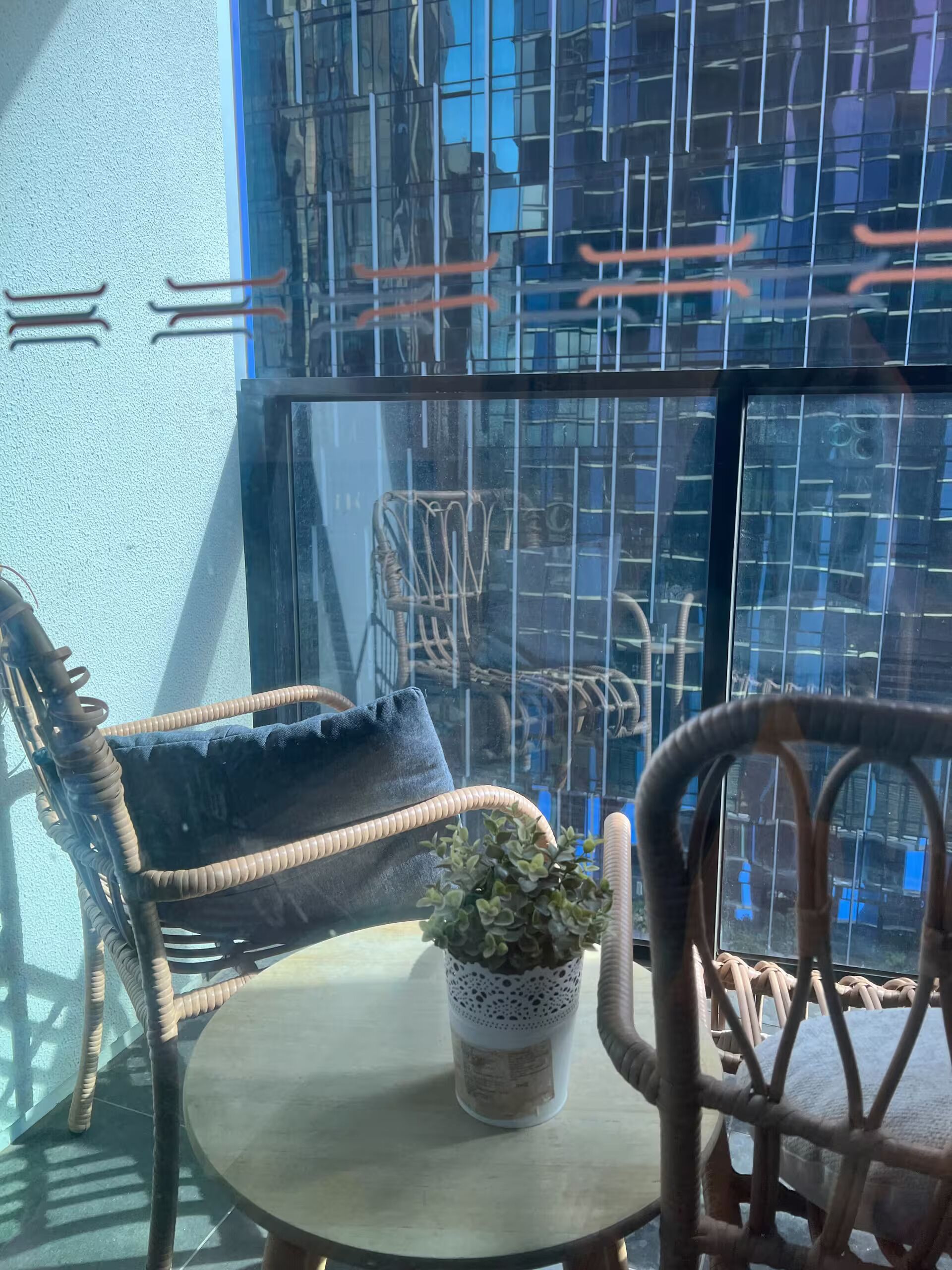 A room with a round table, wicker chairs, a potted plant, and a view of a building through a glass window.