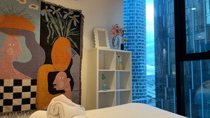 A hotel room with a large window, a bed with a rolled white towel, a white shelving unit, and a colorful tapestry on the wall.