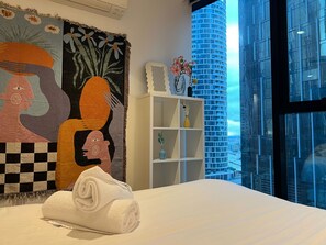 Ein Hotelzimmer mit Bett, Wandteppich, weißem Regal mit Dekoration und Blick auf ein modernes Gebäude.