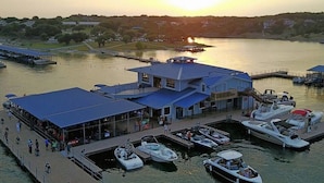 Un quai au bord de l’eau avec des bateaux, un bâtiment doté d’un toit bleu et un coucher de soleil en arrière-plan.