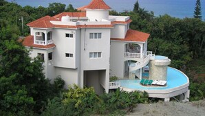A large, multi-story building with a red-tiled roof, surrounded by lush greenery and overlooking a body of water. - Private Ocean View Villa - Chef & Staff Included (Puerto Plata)