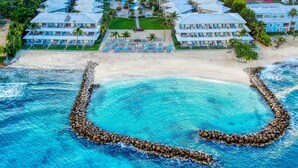 An aerial view of a resort with multiple buildings, a beach, and a clear blue pool with a rock barrier.
