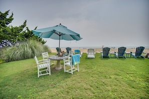 A grassy outdoor seating area with a table and chairs, a large umbrella, and beach chairs lined up facing the ocean. - Chic Virginia Beach Retreat - Pool, Steps to Beach (Virginia Beach)