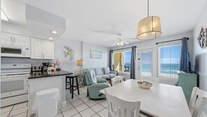 Eine moderne Küche mit weißen Schränken, eine Essecke mit weißem Tisch und Stühlen, ein Deckenventilator und Meerblick durch große Fenster.