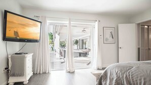 A bedroom with a bed, a TV mounted on the wall, a desk, and a view of the outdoor area through a glass door.