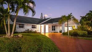 A two-story house with a paved driveway leading to the entrance, surrounded by palm trees and well-maintained lawns.