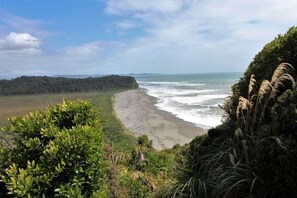 A coastal landscape with a beach, vegetation, and a distant forested area. - Wildside Lodge (romantic off-grid cabin) (Westland District)