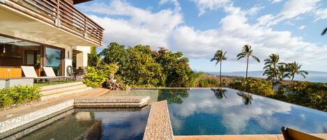 A modern house with a large infinity pool, a wooden deck, and a view of the ocean.