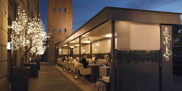 Alfresco dining area with tables and chairs along a covered walkway, surrounded by a brick tower and a tree.