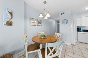 A dining area with a round wooden table, white chairs, a chandelier, and a clock on the wall. - Gulf Shores Beachfront Escape | Sleep 4 | Pool + On-Site Dining | Book Your Stay (Gulf Shores)