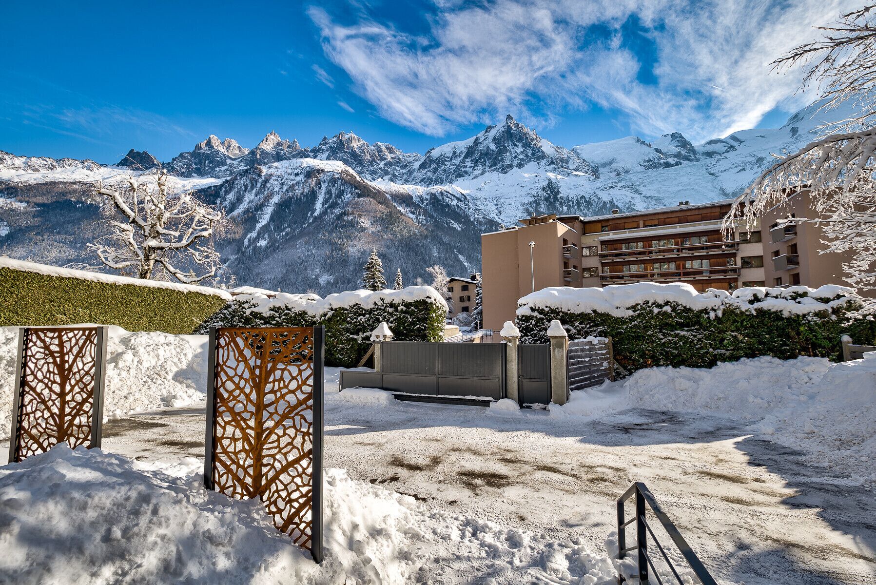 A snow-covered courtyard with a mountain backdrop, a building with balconies, and a wooden fence.