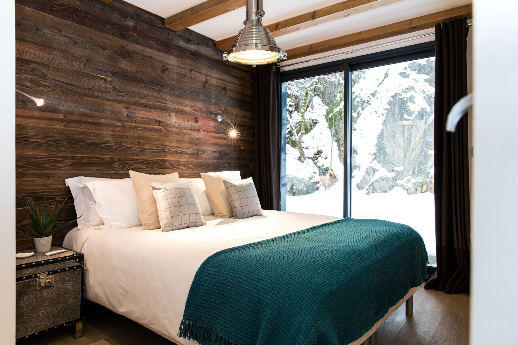 A bedroom with a wooden headboard, a bed with white bedding and a teal throw, a bedside table with a plant, and a large window with a snowy mountain view.