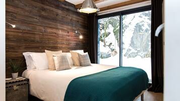 A bedroom with a wooden headboard, a bed with white bedding and a teal throw, a bedside table with a plant, and a large window with a snowy mountain view.