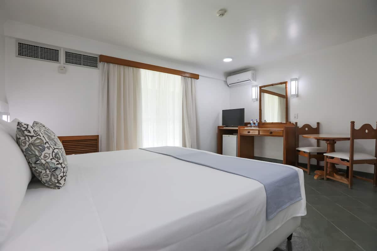 A hotel room with a bed, a desk, a chair, a television, and an air conditioning unit.