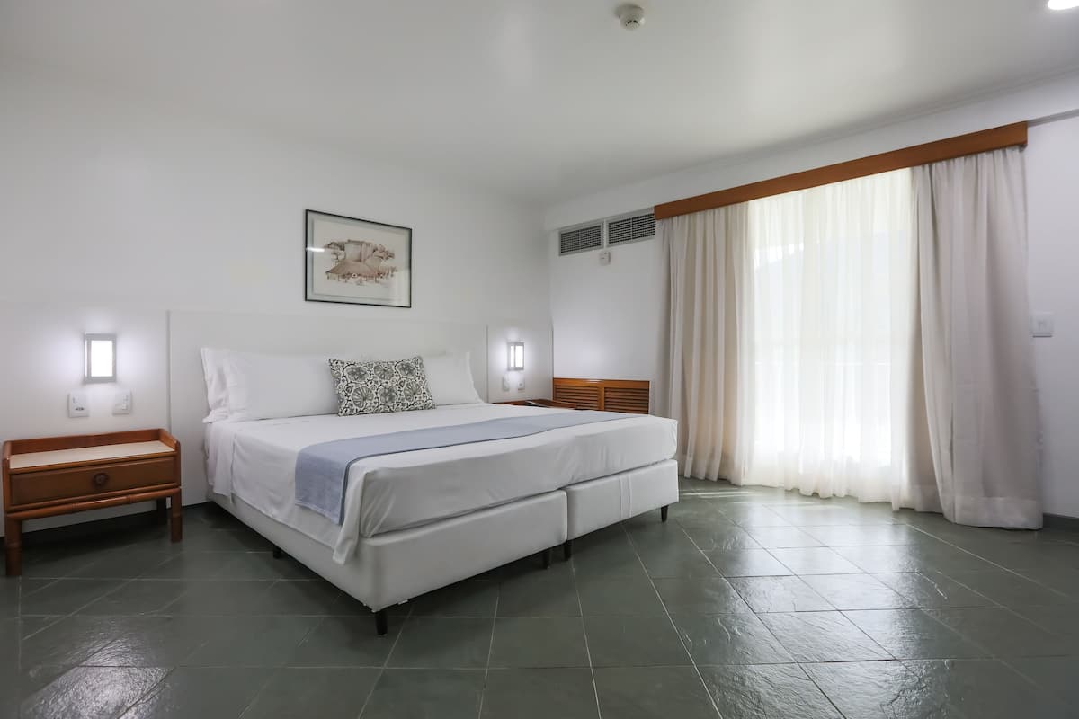 A bedroom with a large bed, two bedside tables, a wall-mounted light, a ceiling fan, and a window with curtains.