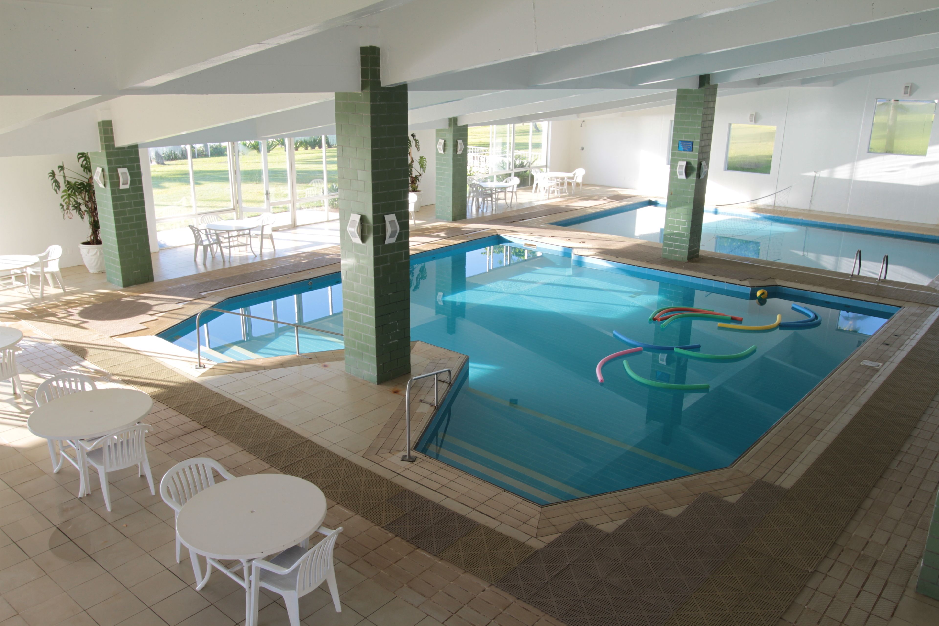Una piscina interior con agua cristalina, rodeada de sillas y mesas blancas, y grandes ventanales con vistas al exterior.
