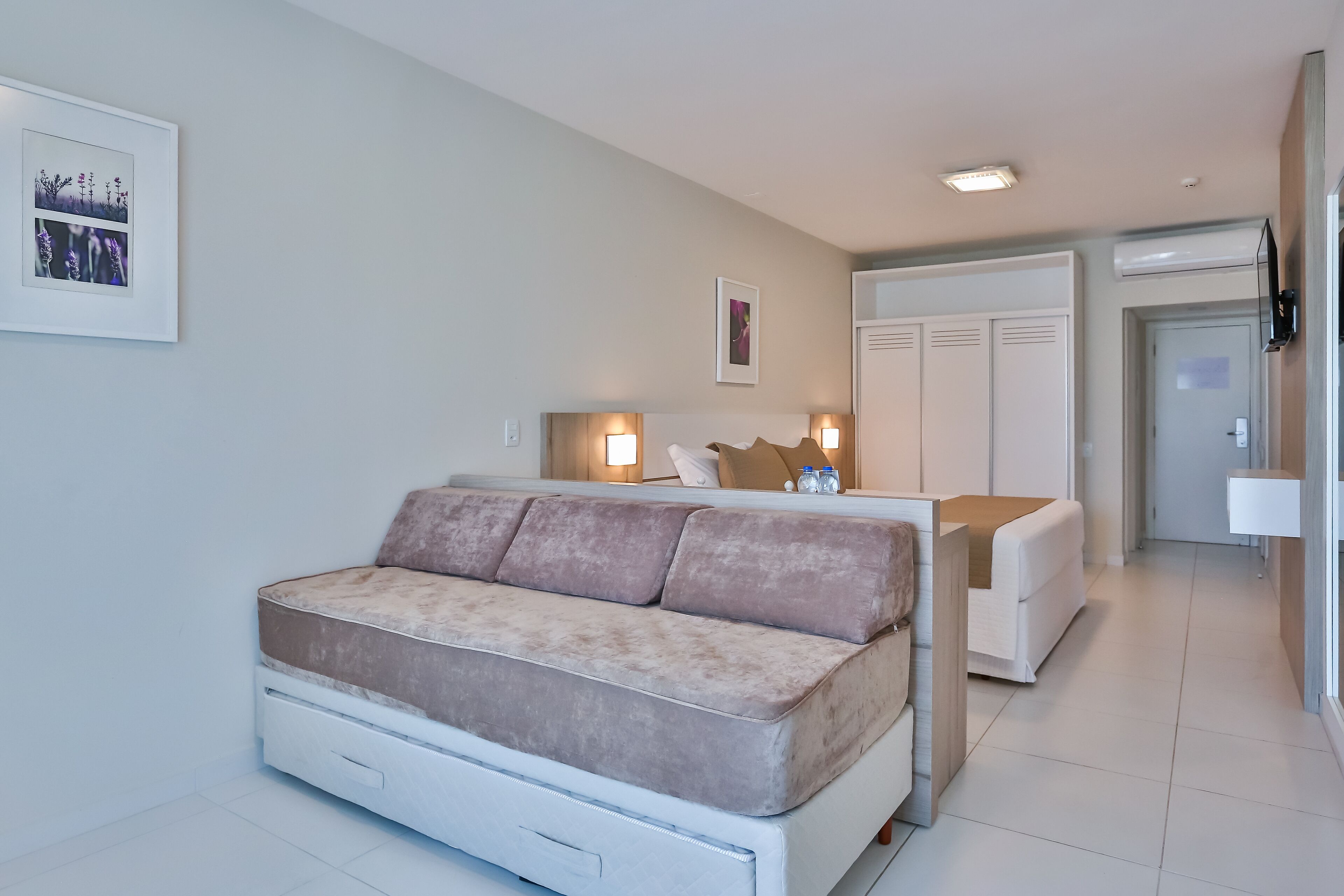 A modern hotel room with a sofa bed, a bedside table, a mirror, a wardrobe, and a framed picture on the wall.
