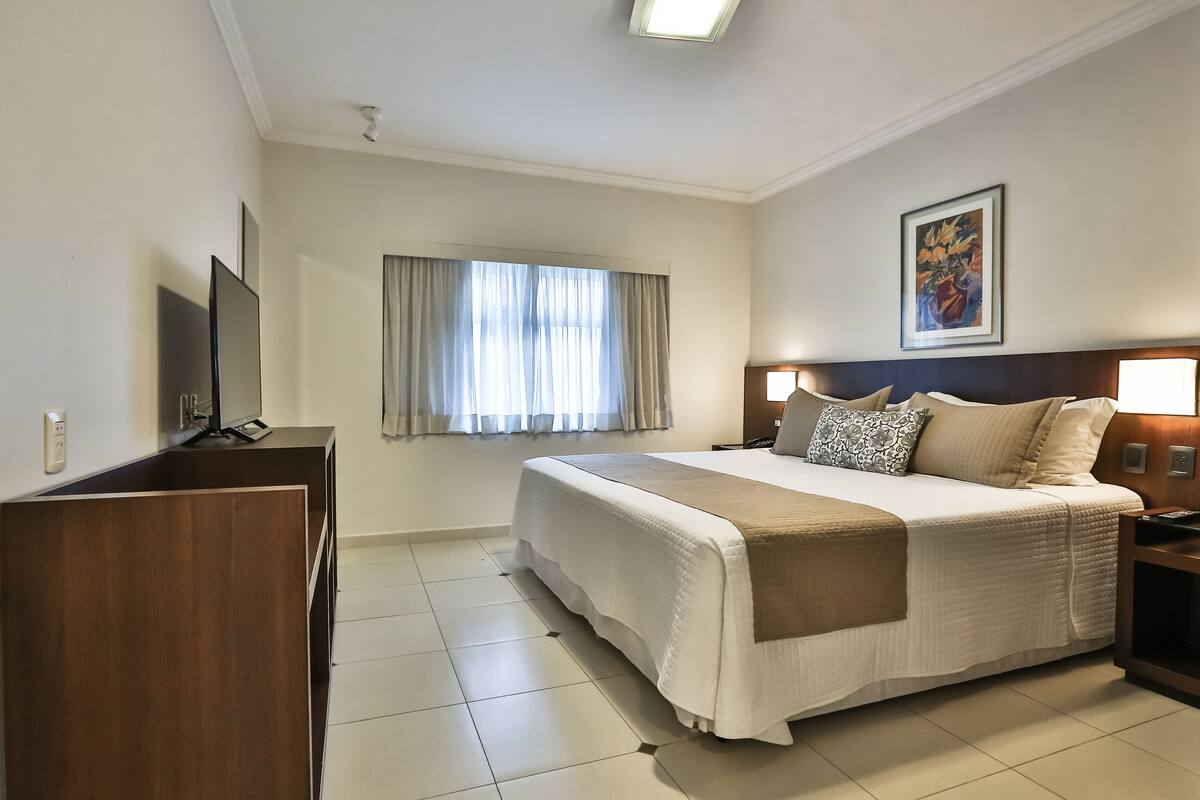 A hotel room with a bed, a TV, a desk, and a window with curtains.