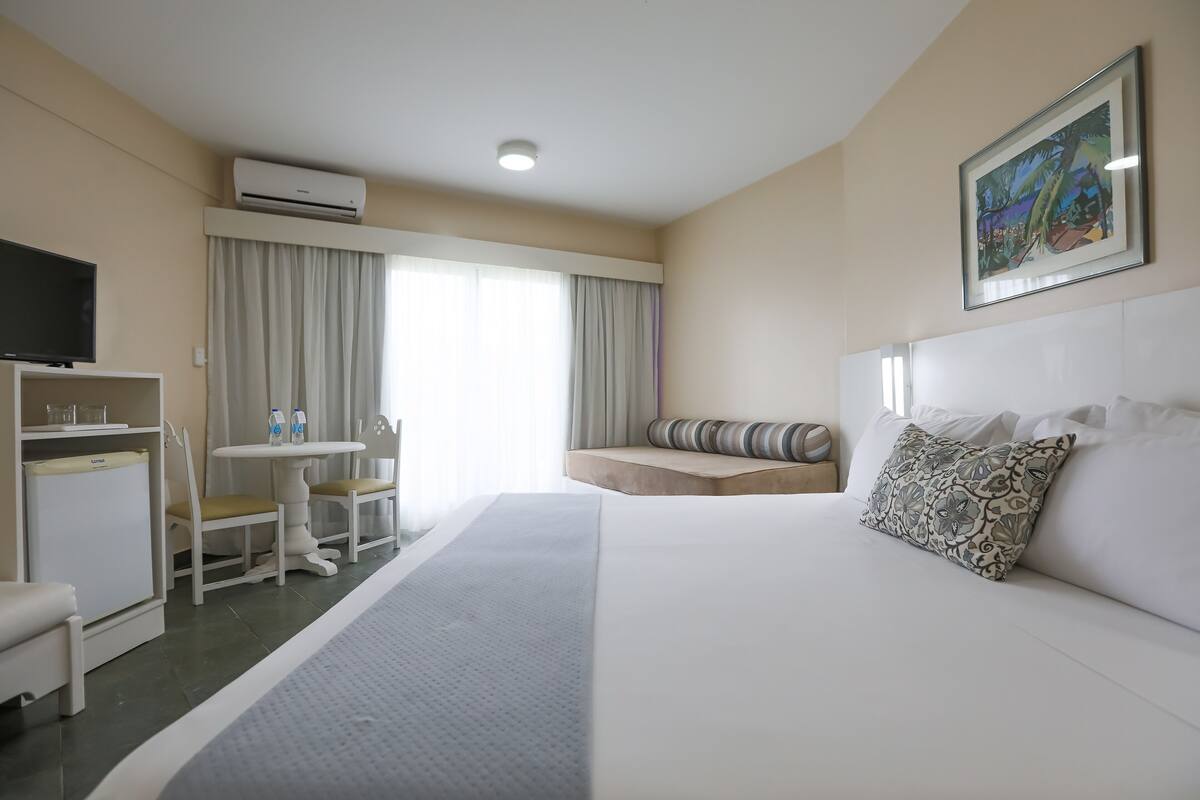 A hotel room with a bed, a TV, a desk, and a window with curtains.