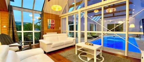 Un interior moderno con amplios ventanales, un sofá blanco, una mesita y una piscina visible a través del vidrio.