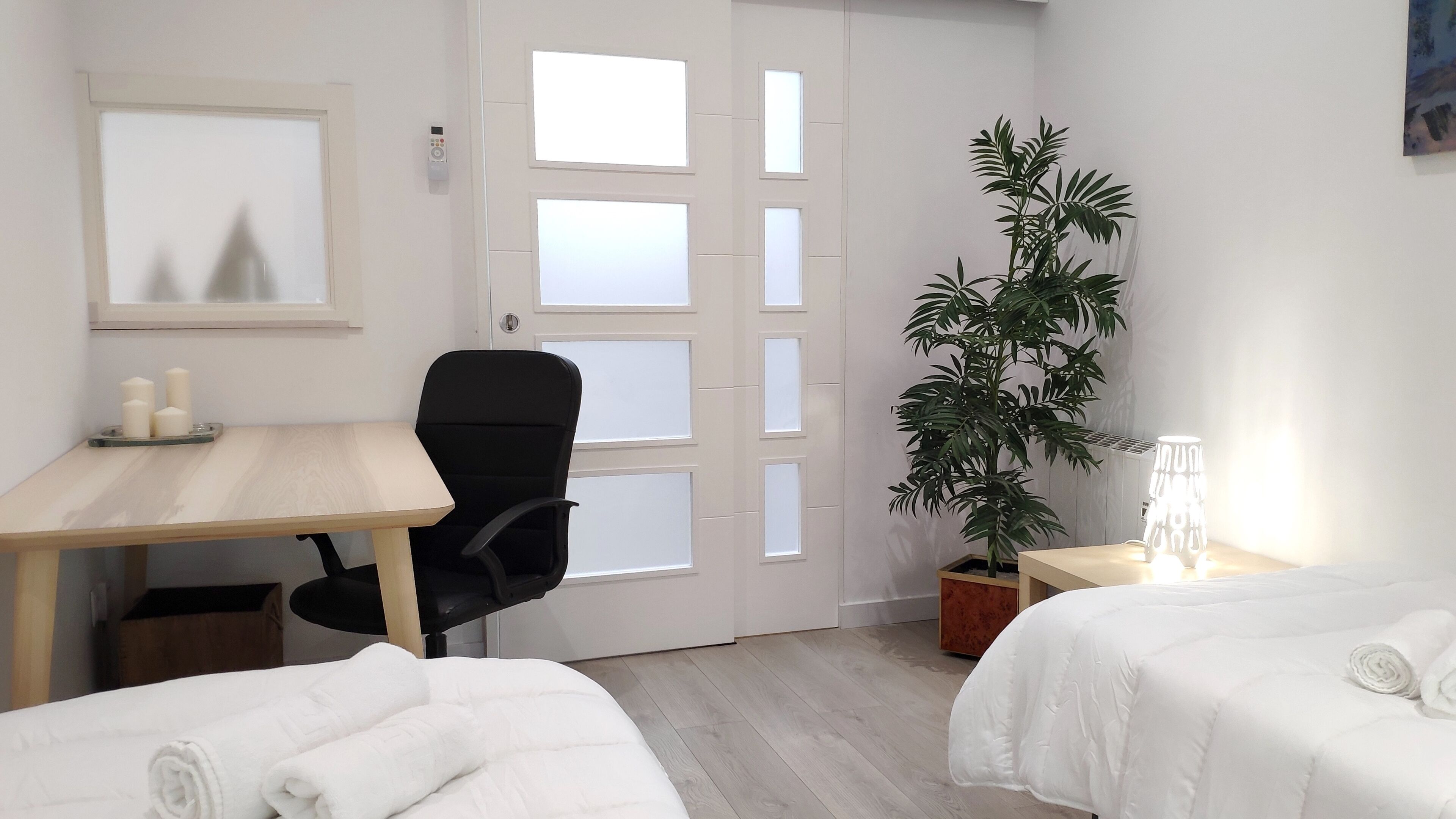 Une chambre d’hôtel avec un bureau en bois, un fauteuil de bureau noir, une plante en pot, une table de chevet avec une lampe et deux lits avec du linge de lit blanc.