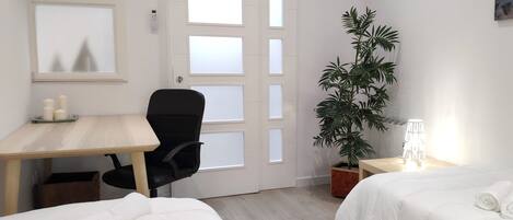 Une chambre d’hôtel avec un bureau en bois, un fauteuil de bureau noir, une plante en pot, une table de chevet avec une lampe et deux lits avec du linge de lit blanc.