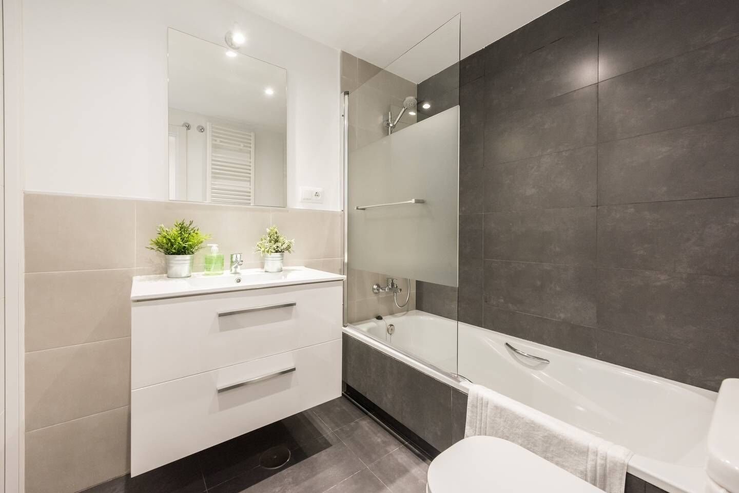 Un baño moderno con tocador blanco, un espejo grande, una bañera y paredes cubiertas de baldosas oscuras.