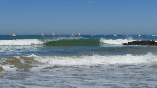 Una playa con olas, costa rocosa y veleros a lo lejos.