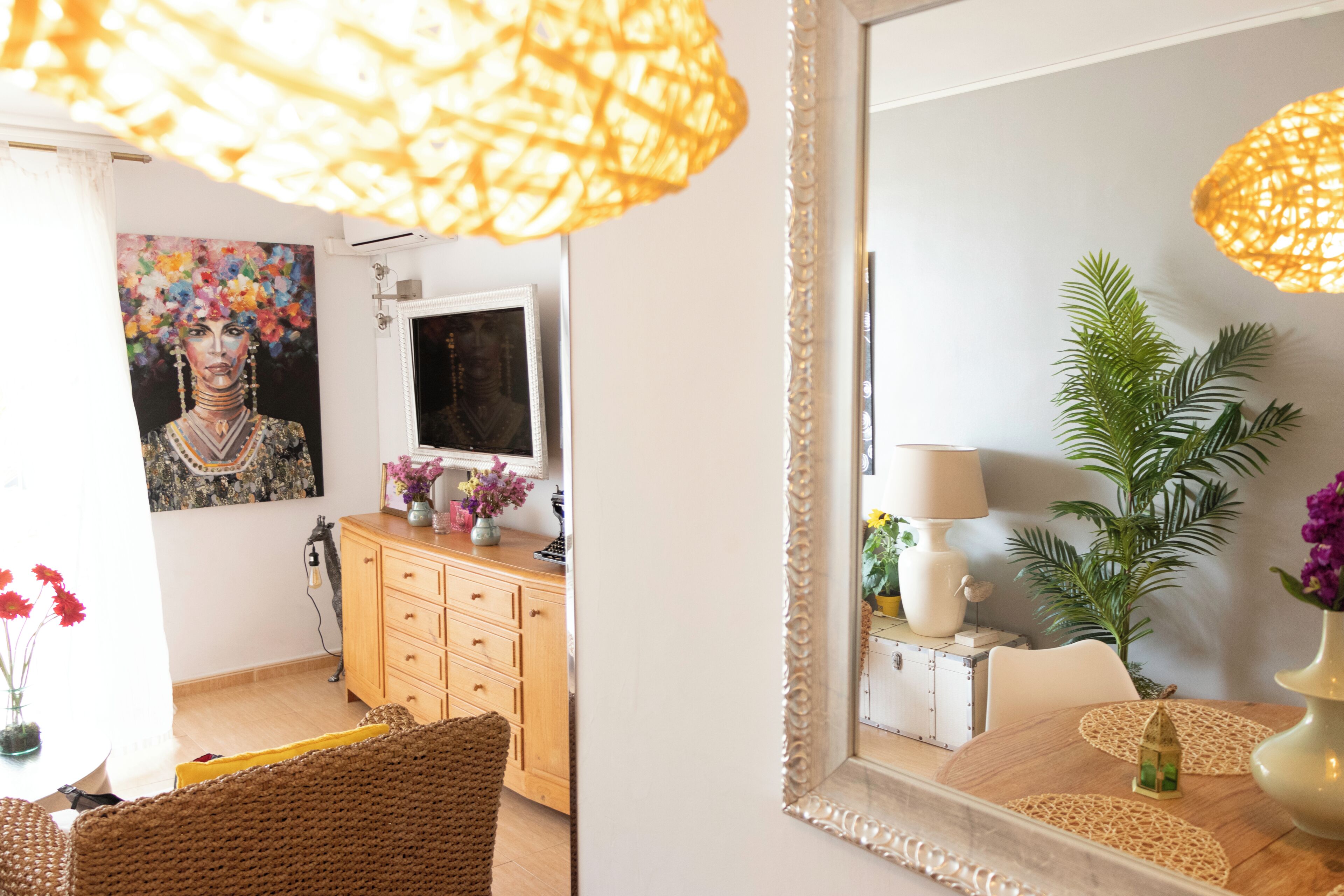 A room with a large mirror, a wooden cabinet, a wicker chair, and a table with a lamp and a vase.