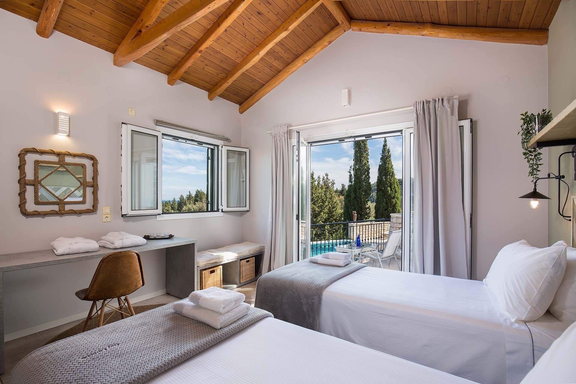 Une chambre d’hôtel avec deux lits, un bureau, une chaise, une grande fenêtre donnant sur la verdure et un plafond avec des poutres en bois.