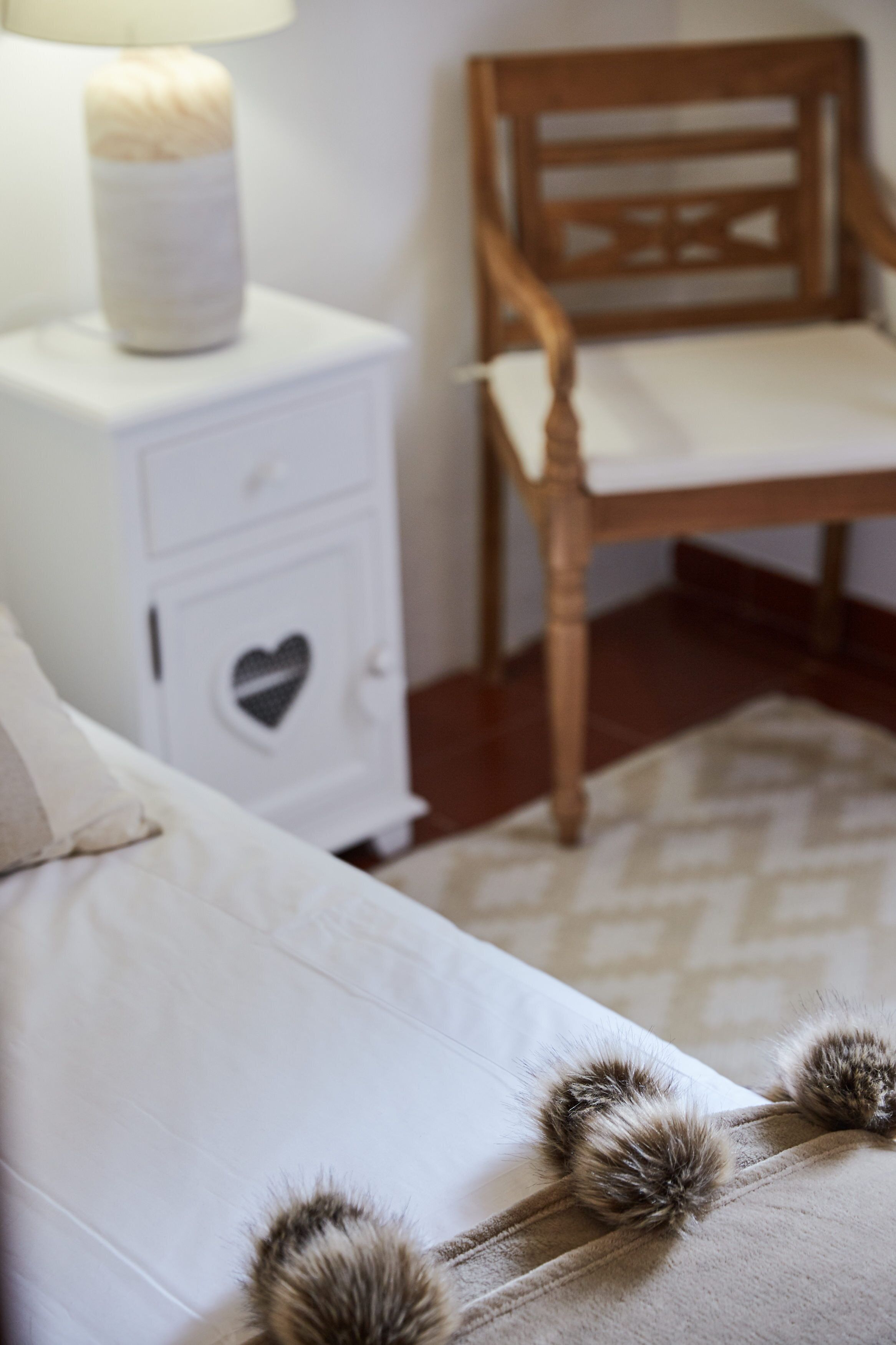 A bedroom with a bed, a nightstand with a heart-shaped drawer, a wooden chair, and a lamp.