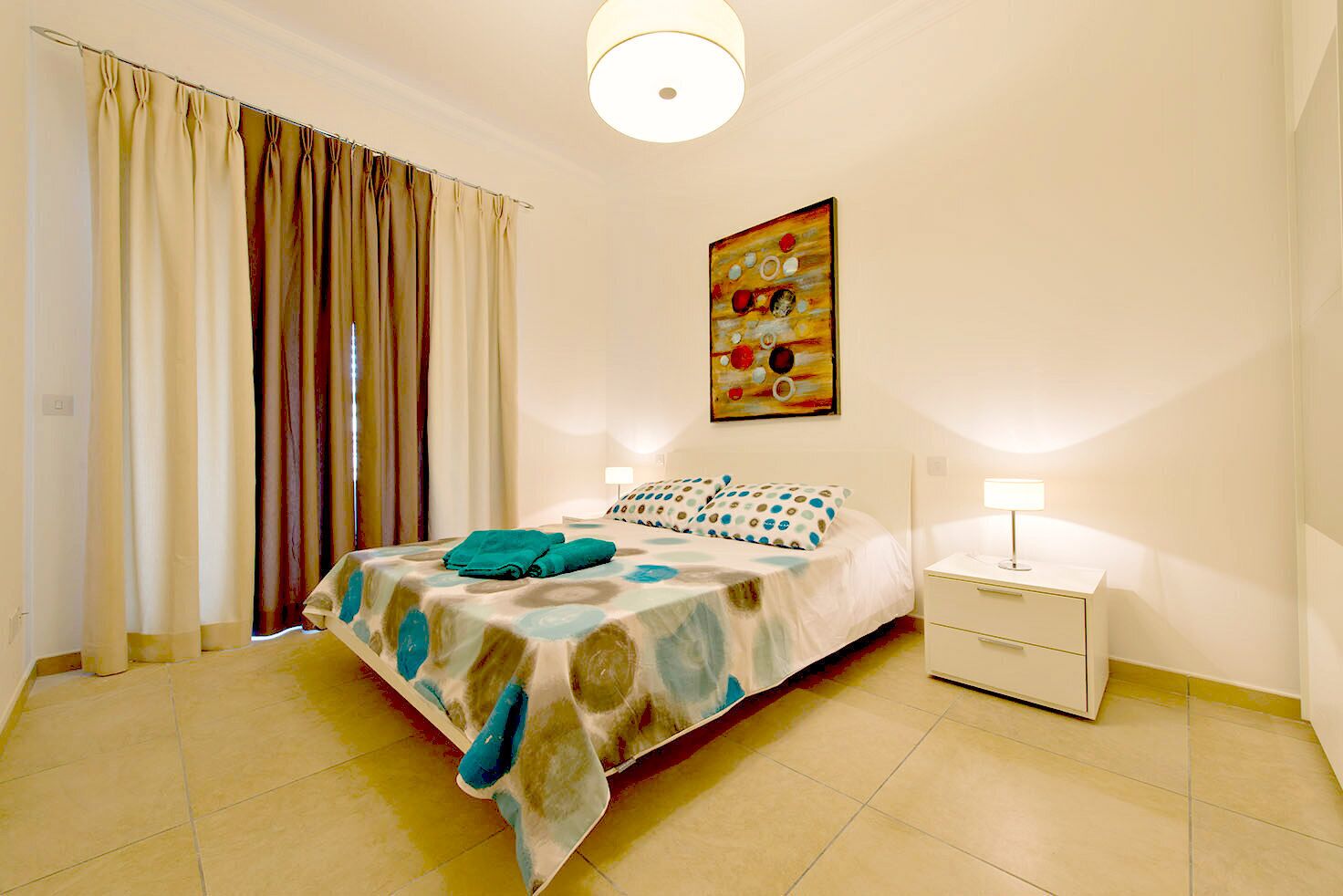 A bedroom with a bed, curtains, a painting, and a nightstand with a lamp.