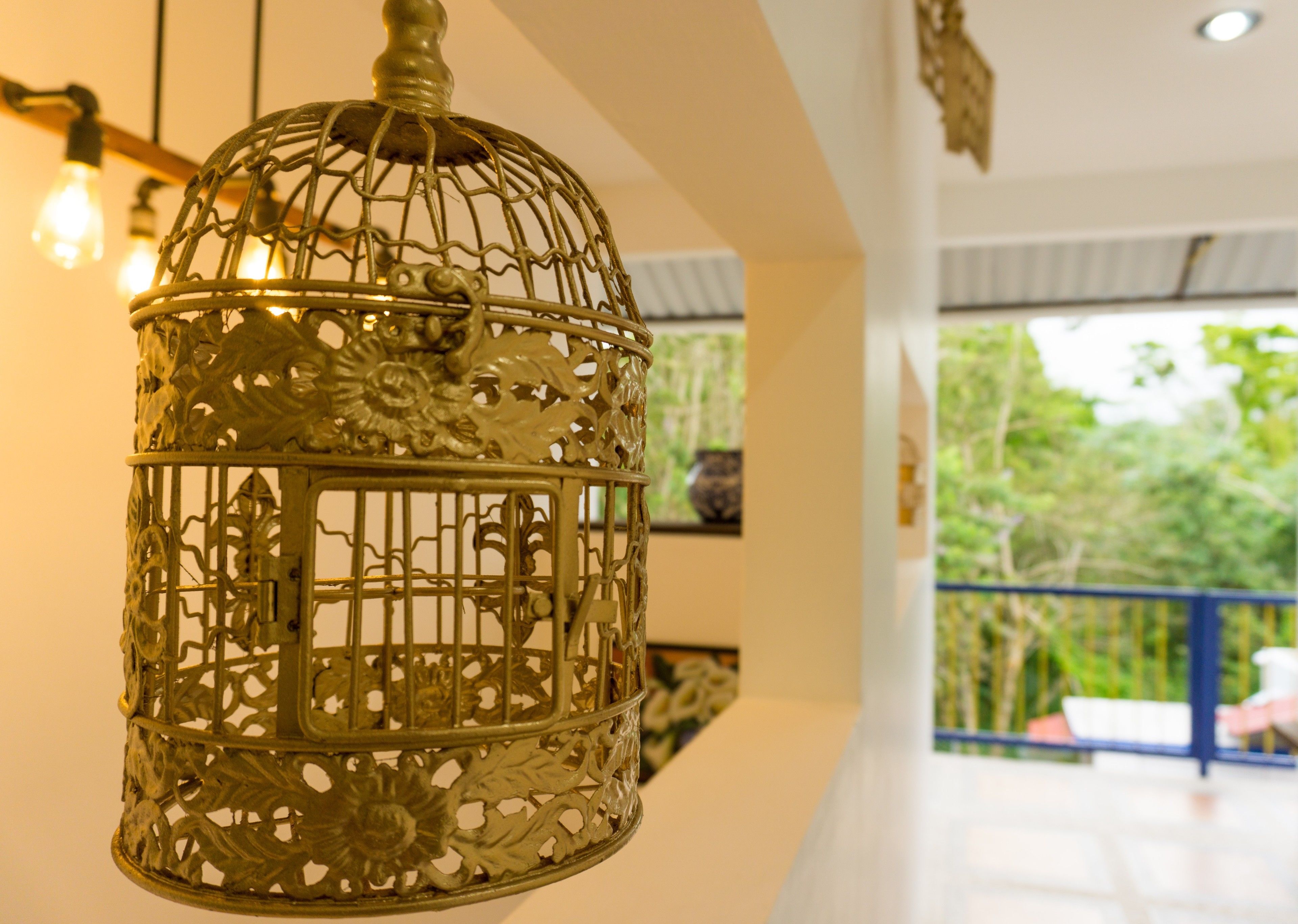 A hanging metal birdcage with intricate designs, a ceiling light, and a view of a balcony with greenery outside.